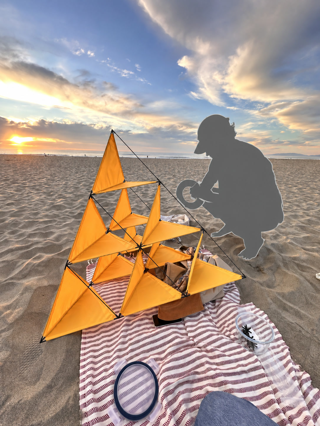 A gray silhouette crouches beside an orange ten-cell tetrahedral kite at Ocean Beach at sunset